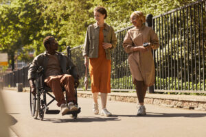 Young multi-ethnic friends walking together along street and talking to disabled man in manual wheelchair