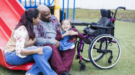Family cuddling disabled child beside wheelchair in play park